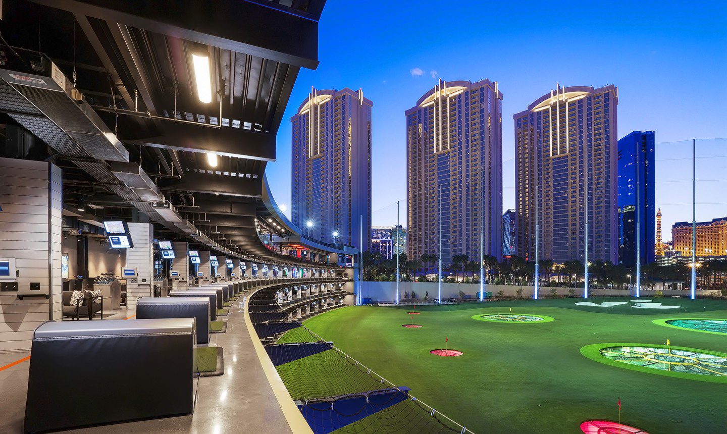 The driving range of a TopGolf venue at night with skyscrapers in the background.