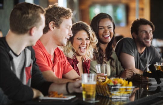 A group of friends smiles and laughs while gathered around a table at Topgolf.