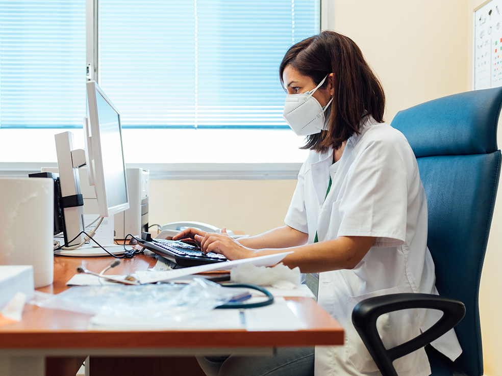 Medical worker wearing a mask sitting at a desk typing on a computer