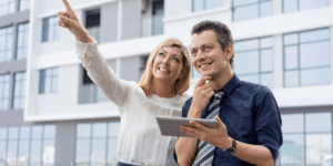 A man and woman smile while the man holds a tablet and the woman points at something in the distance.