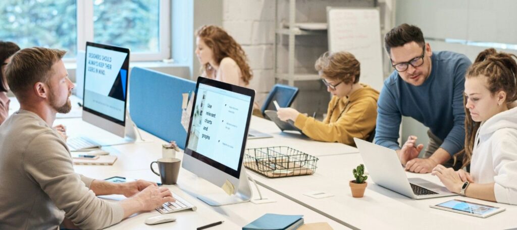 A group of tutors sits with children at a long desk.