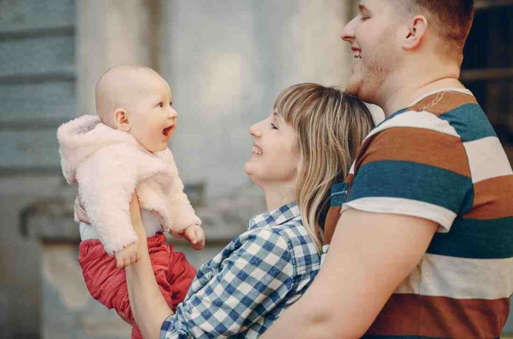 A mother and father smile at a baby who is smiling back at them.