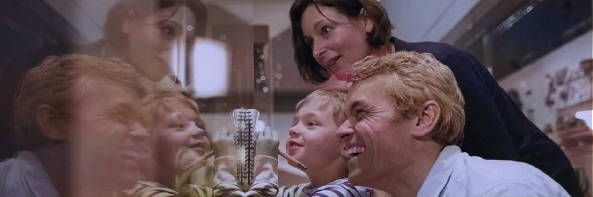A family of three smiles while looking at a reflective wall. A small child writes in a notebook held against the wall.