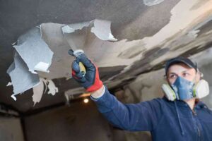 A contractor in protective equipment removes spackle from a wall.