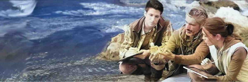 Three people crouch in a shallow body of water, holding clipboards and discussing with each other.