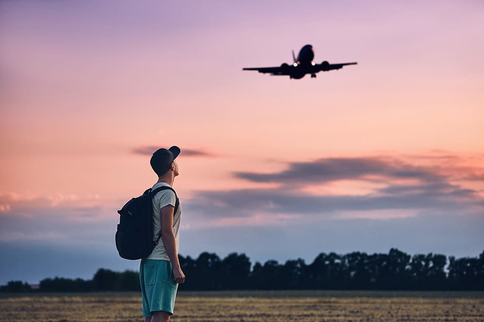 A man stands and looks at a commercial airliner flying overhead.