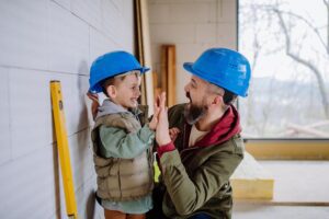 A father & son wearing construction hats smile & high-five each other.