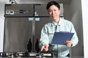 A repairman holding a notebook examines a cooling unit for a refrigerator.
