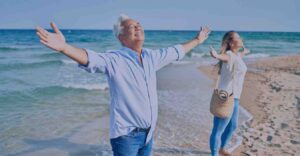A man and a woman stand with their arms out at the beach, smiling and looking towards the sky.