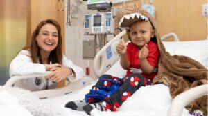 A small child sitting up in a hospital bed wearing a dinosaur blanket smiles. A doctor is crouching next to the bed.