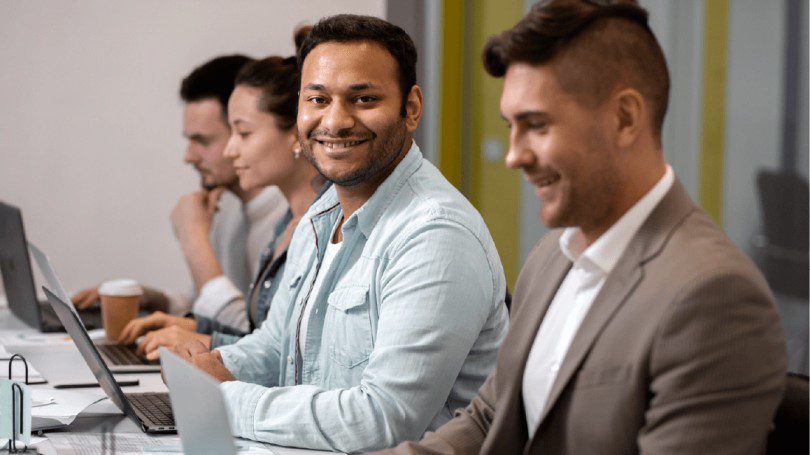 A group of four people in business attire work on their laptops. One man is smiling at the camera.