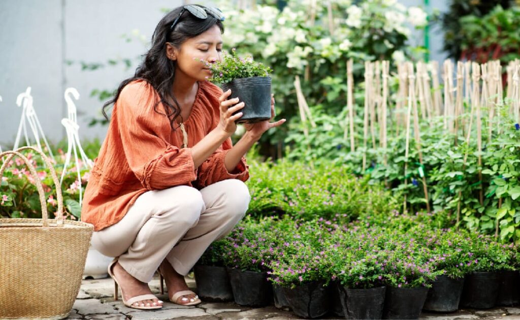 A woman crouches next to a small garden smelling a small pot with a plant in it.