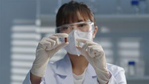 A researcher wearing goggles and a mask compares two different pills in her hands.