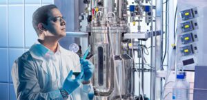 A biologist in a white coat and goggles studies a piece of machinery.