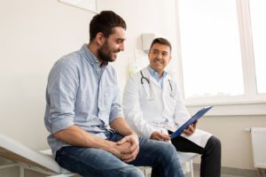 A man and a doctor sit together at a medical office table. The doctor is holding a tablet.