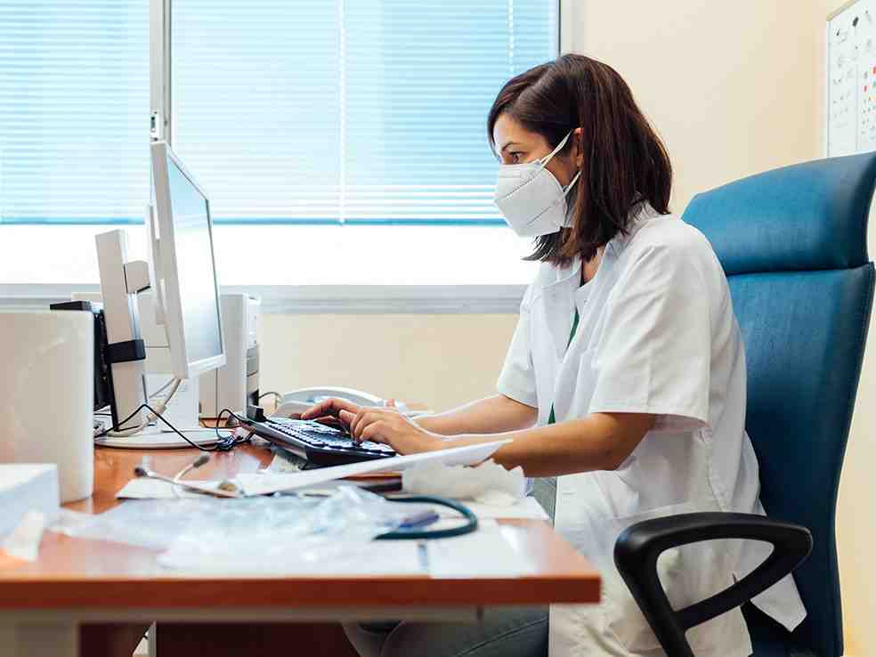 A woman in medical scrubs wearing a mask works on a computer at a desk.