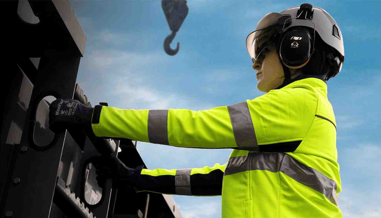A worker in a high-visibility jacket operates machinery, wearing protective gear and focused under a blue sky.