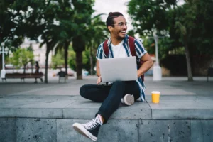 A young man sits on a bench in a tropical campus area, smiling and working on a laptop.