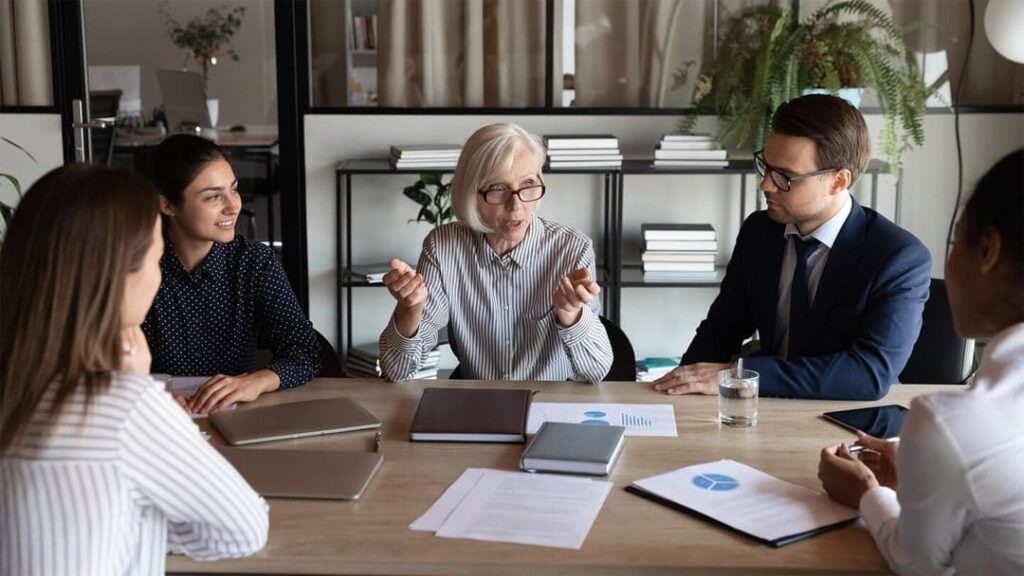An older woman sits at an office table discussing with her team, who are gathered around the table.