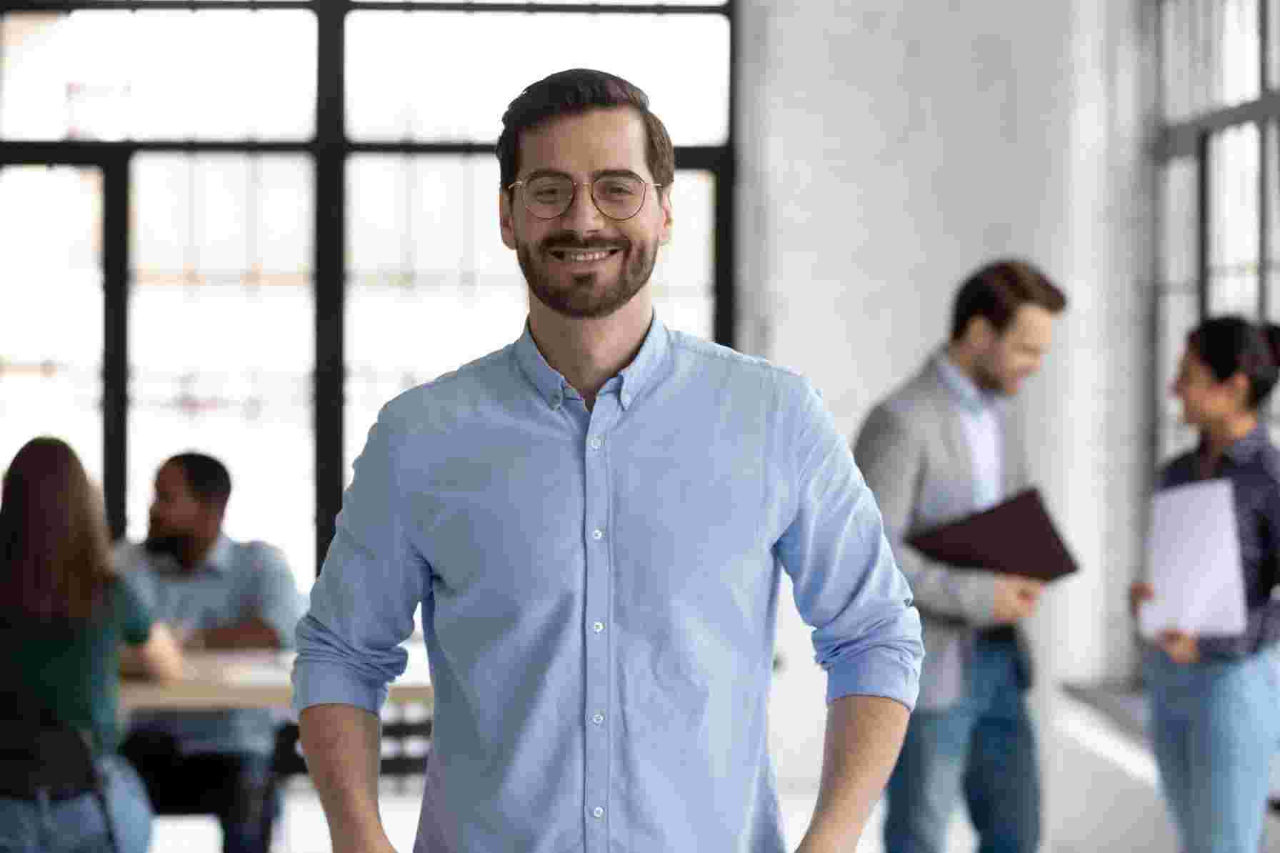 Blog 43 Portrait of happy millennial male business owner in modern office. Businessman wearing glasses, smiling and looking at camera. Busy diverse team working in background. Leadership concept. Head shot.