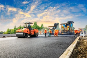 Construction workers and an asphalt paver lay down asphalt on a road. The sky is partly cloudy as the sun sets.