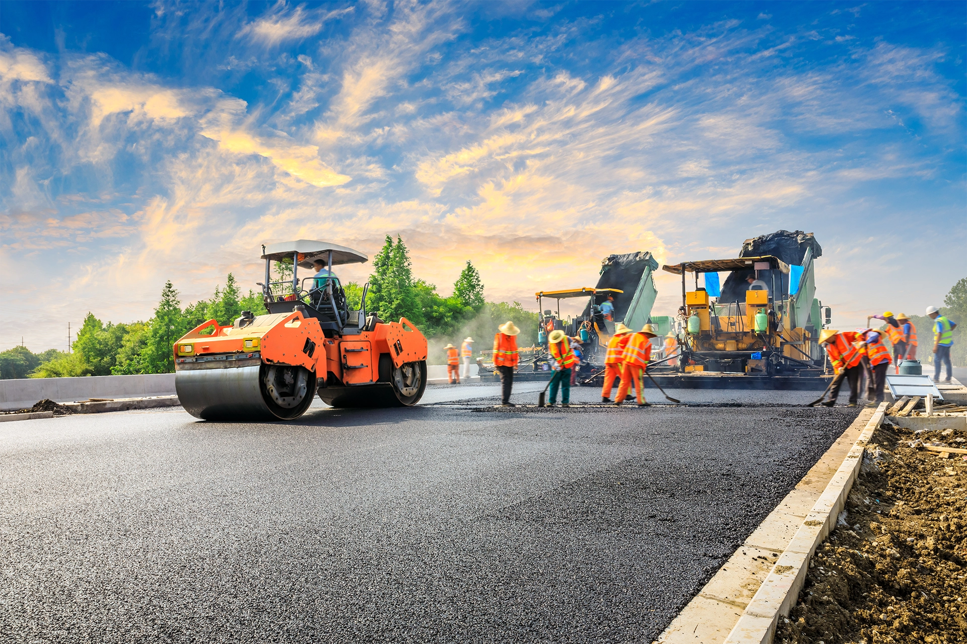 Construction workers and an asphalt paver lay down asphalt on a road. The sky is partly cloudy as the sun sets.
