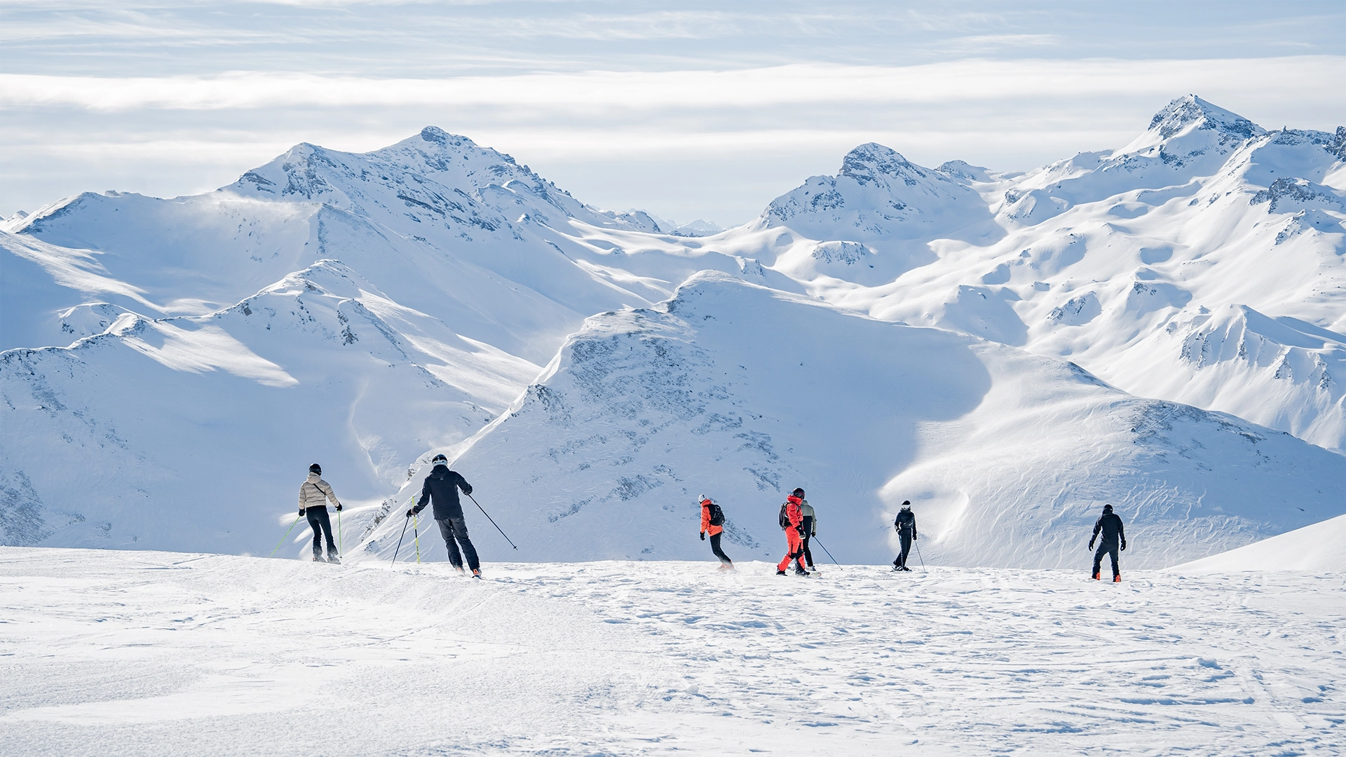 How To Homepage 1 People ride skis down a slope with snow-covered mountains in the background.