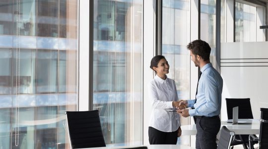 A man and woman in business attire smile and shake hands next to glass windows in an office building.