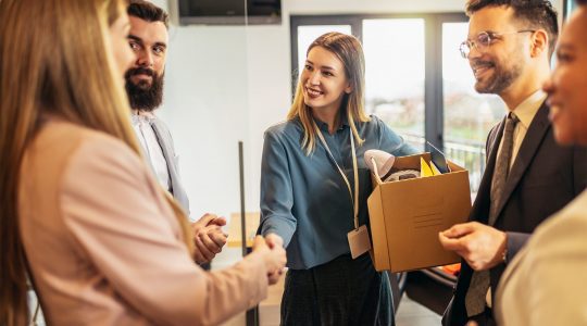 A woman in business attire shakes the hands of coworkers while setting up her desk on her first day at work.