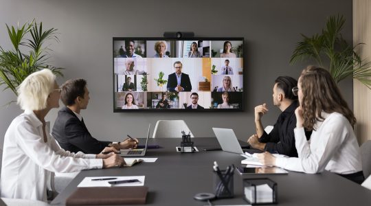 Four people in business attire sit at a conference table, looking at a TV screen during a video meeting with other employees. They are discussing ADP integrations for their HR team.