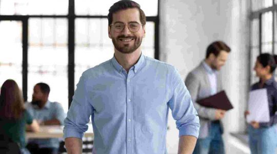 Portrait of happy millennial male business owner in modern office. Businessman wearing glasses, smiling and looking at camera. Busy diverse team working in background. Leadership concept. Head shot.