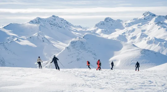People ride skis down a slope with snow-covered mountains in the background.