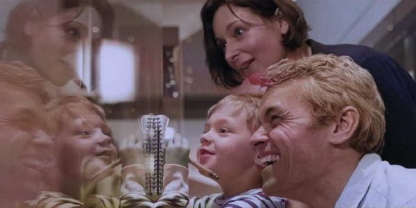 A family of three smiles while looking at a reflective wall. A small child writes in a notebook held against the wall.