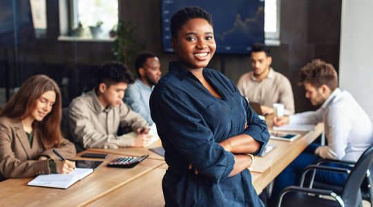 An office meeting room with employees having discussions and their manager standing in a blue shirt. Her organization uses Hire2retire for Oracle to Okta Directory Integration.