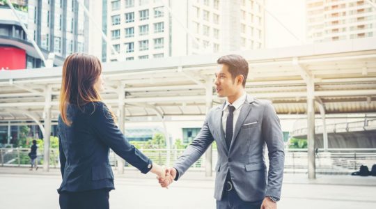 A man and a woman wearing suits shake hands at a train station.
