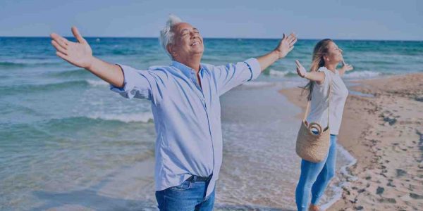 A man and a woman stand with their arms out at the beach, smiling and looking towards the sky.