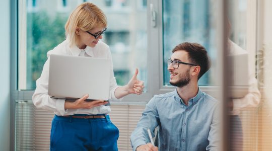 An office manager standing in a white shirt giving feedback to a young intern showing thumbs up. Her organization uses Hire2retire for UKG Pro to Google Workspace